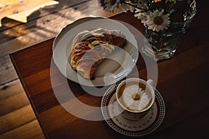 Cup of cappuccino coffee and croissant / brioche on a wooden table with freshly gathered daisy flowers. Italian breakfast.