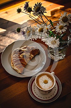 Cup of cappuccino coffee and croissant / brioche on a wooden table with freshly gathered daisy flowers. Italian breakfast.