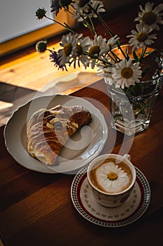 Cup of cappuccino coffee and croissant / brioche on a wooden table with freshly gathered daisy flowers. Italian breakfast.