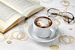 Cup of cappuccino coffee, reading glasses and book on white table with traces of coffee cups