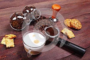 Cup of cappuccino, crackers, cookies, holder with ground coffee, tamper and cans of coffee beans on wooden table