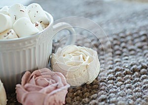 A cup of cocoa or chocolate next to a rose-shaped marshmallow. Warm cozy square background