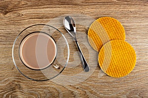 Cup of cocoa with milk in saucer, spoon, two wafers on wooden table. Top view