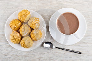 Cup of cocoa with milk on saucer, teaspoon, profiterole in plate