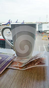 Cup of coffee in airport. Passport and money on the table. Planes, buses and jet bridge blurred on background