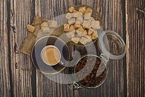 A Cup of coffee, coffee beans in a glass jar and pieces of brown sugar on a wooden background
