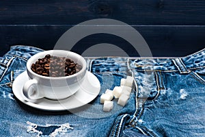 Cup with coffee beans, refined sugar lay on jeans near zip, denim background. Fresh brewed coffee concept. Mug full of