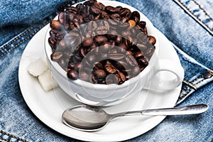 Cup with coffee beans, refined sugar and spoon on plate, denim background. Freshly roasted coffee concept. Mug full of