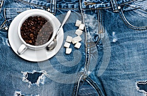 Cup with coffee beans, spoon and refined sugar lay on jeans near zip, denim background. Fresh brewed coffee concept. Mug