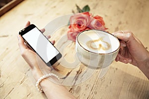 Cup of coffee in beautiful woman hands. Lady using mobile phone internet in cafe. Blank screen for layout. Red roses flowers behin photo