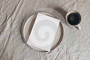 Cup of coffee and blank greeting card, invitation on ceramic plate. Moody breakfast table mockup scene. Beige linen