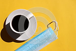 Cup of coffee and blue protective pandemic face mask on yellow background. Top view, copy space. Hard light, shadow.