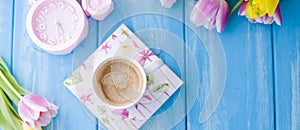 A cup of coffee on a blue wooden background. Bright colors. Bouquet of flowers yellow and pink. The pink clock is like a bicycle.