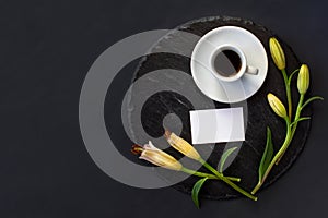 Cup of coffee and buds of lily flowers with clean white business card without massage on a round slate on a black background