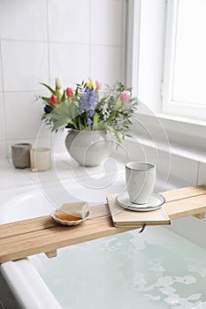 Cup of coffee, candles, book and soap on wooden board. Elegant Scandinavian bathroom interior with white tiles.Blurred