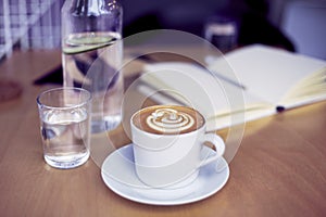 Cup of coffee cappuccino, glass of pure water, bottle on wooden table, bright interior daylight