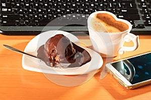 Cup of coffee and chocolate cake next to computer.