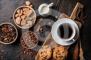 Cup of coffee with chocolate drop cookies, bowls of brown cane lump sugar, crystal sugar sticks, milk and coffee beans