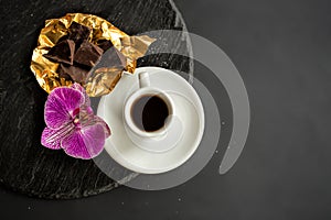 A cup of coffee, chocolate in gold foil and a orchid on a black slate on a black background.