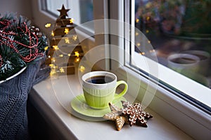 Cup of coffee and Christmas cookie near window in daylight with festive decoration and warm lights on background.