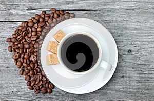 A cup of coffee with coffee beans and cane sugar on a wooden background. White coffee cup and saucer
