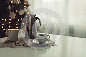 A cup of coffee and a coffee pot with a candlestick stand on the table against the background of a New Year tree