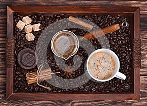 Cup of coffee, coffeepot, biscuit, cinnamon, anise, sugar, coffee beans on a wooden tray.