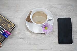 Cup of coffee with cookies, purple daisy flower and notebook with pen and black phone on white wooden table background.