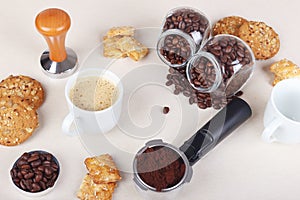 Cup of coffee, crackers, cookies, holder with ground coffee, tamper and cans of coffee beans on table