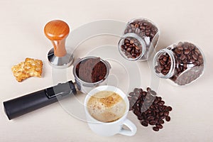 Cup of coffee, crackers, holder with ground coffee, tamper and cans of coffee beans on table