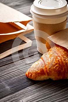 Cup coffee and croissant in paper bag on wooden background