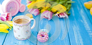 A cup of coffee and donuts on a blue wooden background. Bouquet of flowers yellow and pink. The pink clock is like a bicycle.
