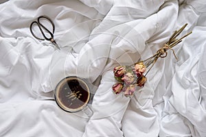 Cup of coffee, dry roses and vintage scissors on bed