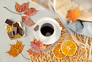 Cup of coffee on a gray background with autumn leaves, a warm cloak, a wicker napkin, pieces of chocolate and slices of dried