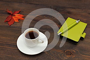 Cup of coffee, green pocketbook, pen and autumn red leaf on wooden table