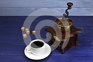 A cup of coffee and coffee grinder on a blue wooden background. White coffee cup and saucer. Copy space. Place for text