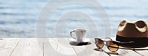 Cup of coffee, hat and sunglasses on a wooden table with beautiful sea view at the background. Panoramic banner, copy space