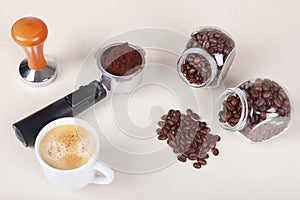 Cup of coffee, holder with ground coffee, tamper and cans of coffee beans on table