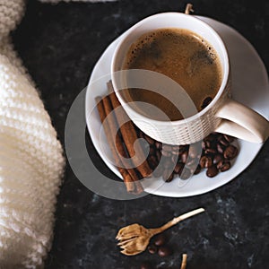 A cup of coffee is the key to a good mood. On a black, dark textural background, a composition of white, cream scarf and a white