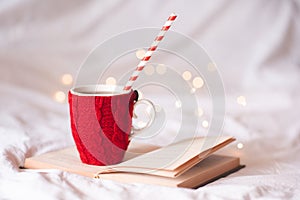 Cup of coffee with knitted red cloth staying on paper bookin bed closeup over glowing lights. Winter holiday season. Cozy