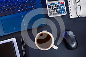 Cup of coffee, laptop with mouse, notebook with pen, calculator, smartphone and glasses on a dark desktop. Business concept