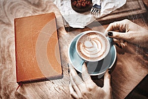 Cup of coffee latte and red book on wooden table or background in woman hands from above