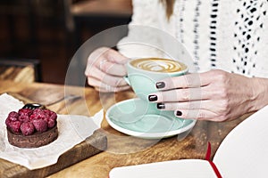 Cup of coffee latte on wooden table or background in woman hands from above. Having lunch in cafe. Opened notebook