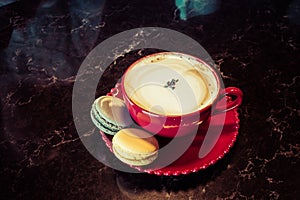 Cup of coffee with macaroon on table. Red cup of cappuccino and saucer with macaroon cookie on desk