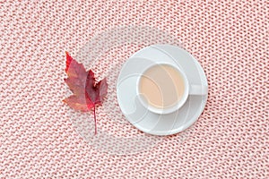 Cup of coffee with milk and red autumn leaf on pastel pink knitted plaid background. Autumn cozy concept. Flat lay, top view, copy