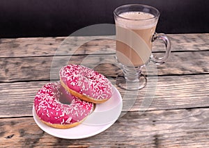 A cup of coffee with milk and two pink donuts standing on wooden textured background