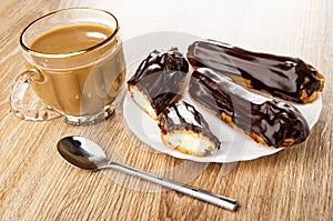 Cup of coffee with milk, whole eclairs and halves of eclair with chocolate in plate, teaspoon on wooden table