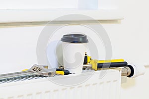 Cup of coffee near plumber tools on heating radiator