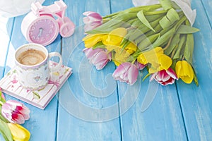 A cup of coffee on a notebook on a blue wooden background. Bright colors. Bouquet of flowers yellow and pink. The pink clock is