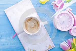 A cup of coffee and a notebook on a blue wooden background. Bright colors. Bouquet of flowers yellow and pink. The pink clock is